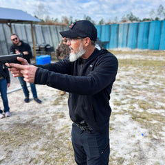 Man with gray beard wearing black Capdre hat and glasses aiming handgun at outdoor shooting range