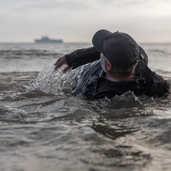 A person in a wet suit and a GORUCK Performance TAC Hat - TOUGHDRY navigates through choppy water with their back to the camera, as a large ship looms on the horizon under a cloudy sky.