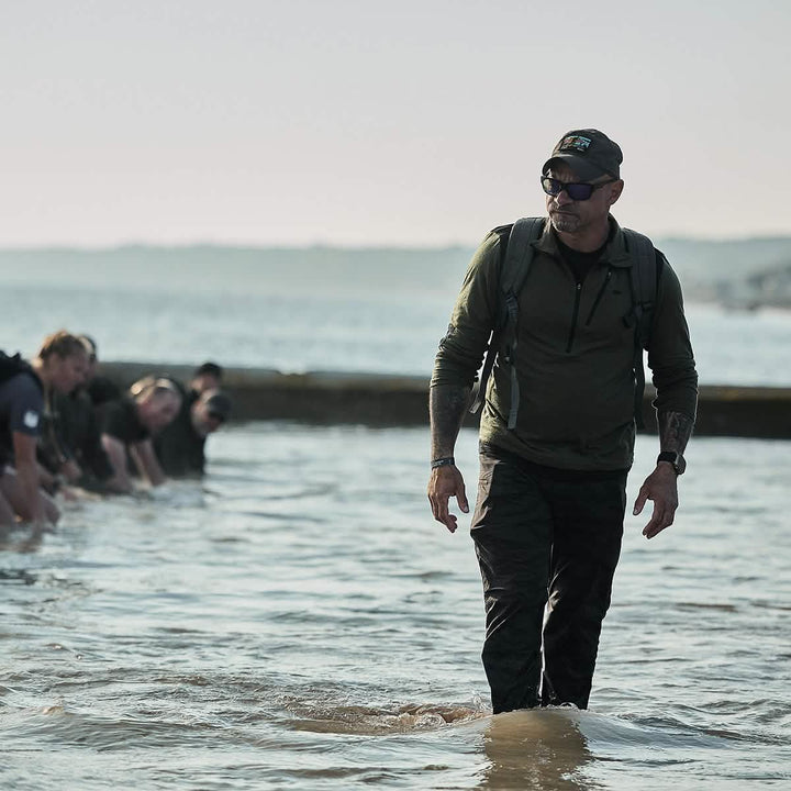 A man in a GORUCK Performance TAC Hat - TOUGHDRY and sunglasses walks through shallow water with a group of people behind him. The team seems to be participating in an exercise or training session at the beach. An overcast sky looms overhead, while the shoreline provides a scenic backdrop.