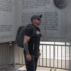A person wearing a black t-shirt, the GORUCK Performance TAC Hat made with TOUGHDRY fabric, and sunglasses stands in front of a stone memorial wall with engraved text. They are carrying a large backpack made from ToughDry fabric and seem to be intently observing the inscriptions.