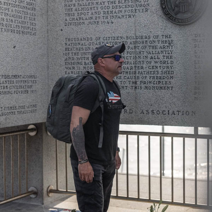 A person wearing a black t-shirt, the GORUCK Performance TAC Hat made with TOUGHDRY fabric, and sunglasses stands in front of a stone memorial wall with engraved text. They are carrying a large backpack made from ToughDry fabric and seem to be intently observing the inscriptions.