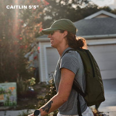 A woman walks outside wearing a green cap, sunglasses, and the Basic Rucker® backpack; text reads "CAITLIN 5'5"".
