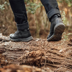 Person wearing black rugged boots and rolled-up pants walking on fallen tree trunk in forest, outdoor gear