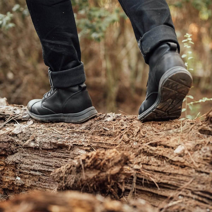 A person wearing the GORUCK MACV-1 Traveler x Carryology - Berserker Viking boots and black pants strides confidently across a fallen tree trunk in a forest, with a backdrop of blurred greenery.