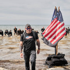 A person wearing a black GORUCK T-shirt and a Performance TAC Hat - TOUGHDRY strolls along a sandy beach. American flags on poles are visible to the right, while several others in wet suits emerge from the water behind them. The sky is cloudy.