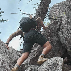Person rock climbing outdoors wearing black GORUCK shorts, tan hiking boots, and carrying a large backpack