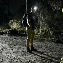 Man wearing GORUCK gear with backpack and headlamp standing on dirt trail at night in forest