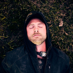 A man in an Indestructible Challenge Windbreaker and cap lies on the grass with his eyes closed, appearing to rest or sleep outdoors.