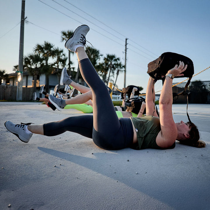 While exercising outdoors under palm trees and power lines, people lay on their backs, holding heavy bags with extended legs. They're sporting Women's Cropped Training Leggings made from ToughFlex fabric, perfect for this squat-proof workout session.