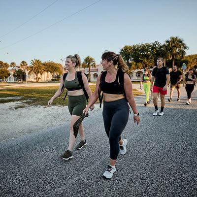A group of people strolls and chats on a sunny road, framed by palm trees and buildings. The women wear Women's Cropped Training Leggings - ToughFlex, offering comfort and flexibility under the warm sun.