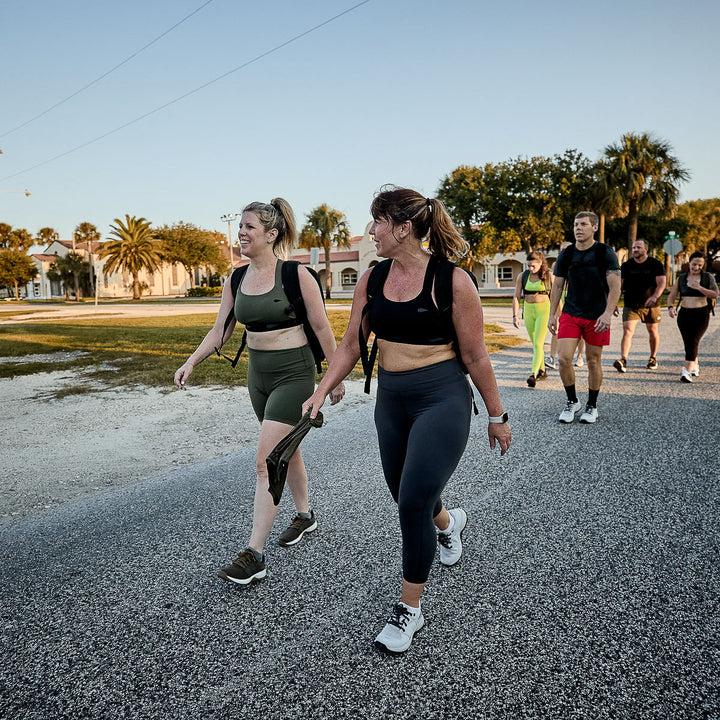 A group of people strolls and chats on a sunny road, framed by palm trees and buildings. The women wear Women's Cropped Training Leggings - ToughFlex, offering comfort and flexibility under the warm sun.