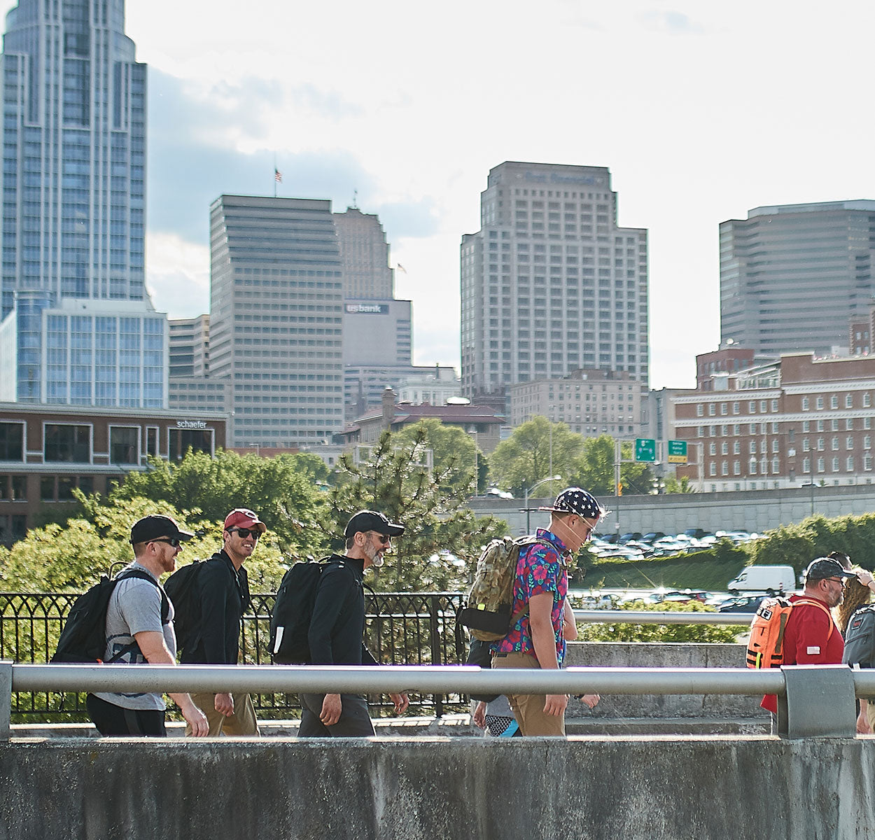 Five people with backpacks walk near a railing in a city, with tall buildings in the background.