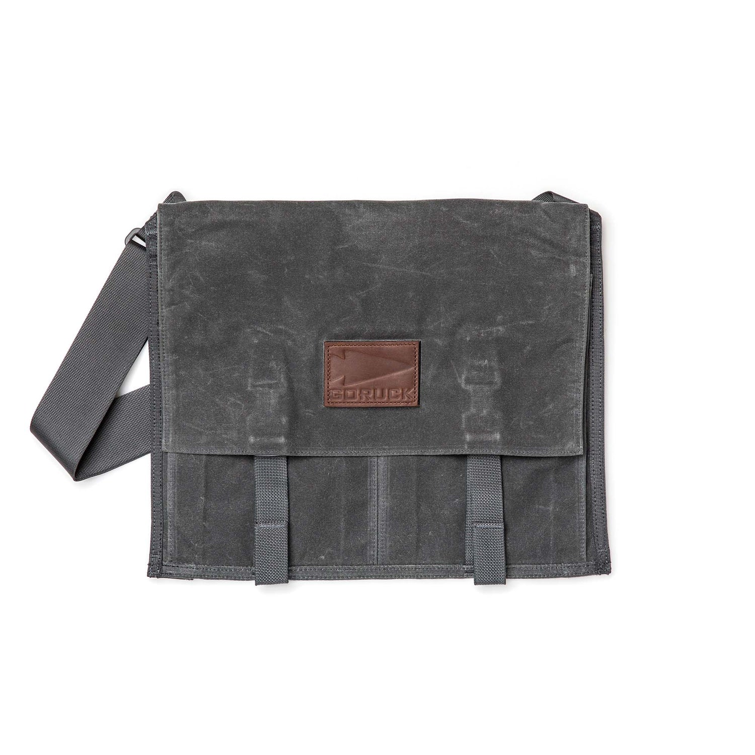 The Waxed Canvas Claymore Sling Bag features a dark gray waxed canvas, a shoulder strap, and a brown leather patch on the front flap for a modern, versatile style.