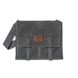 The Waxed Canvas Claymore Sling Bag features a dark gray waxed canvas, a shoulder strap, and a brown leather patch on the front flap for a modern, versatile style.