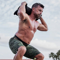 fit man in camo shorts performing weighted squats outdoors with GORUCK sandbag