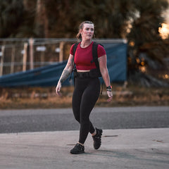 At sunset, a woman in athletic wear and a weighted vest enjoys rucking outdoors on a paved path, comfortably supported by the Padded Hip Belt.