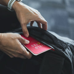 Hands placing a red Field Notes notebook into the zippered pocket of a black GR3 - X-PAC carry-on backpack.