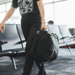 A person in black clothes carries the GR3 - X-PAC, a waterproof black duffel bag, while walking through an airport seating area.