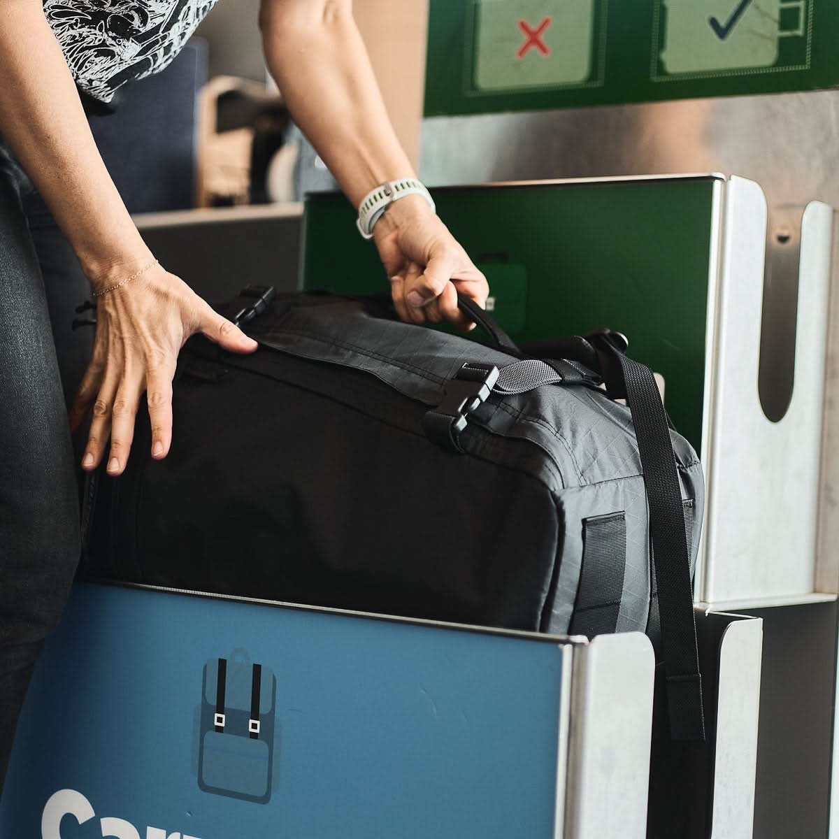 A traveler places the GR3 - X-PAC, a black waterproof carry-on backpack, into a luggage sizer at airport check-in.