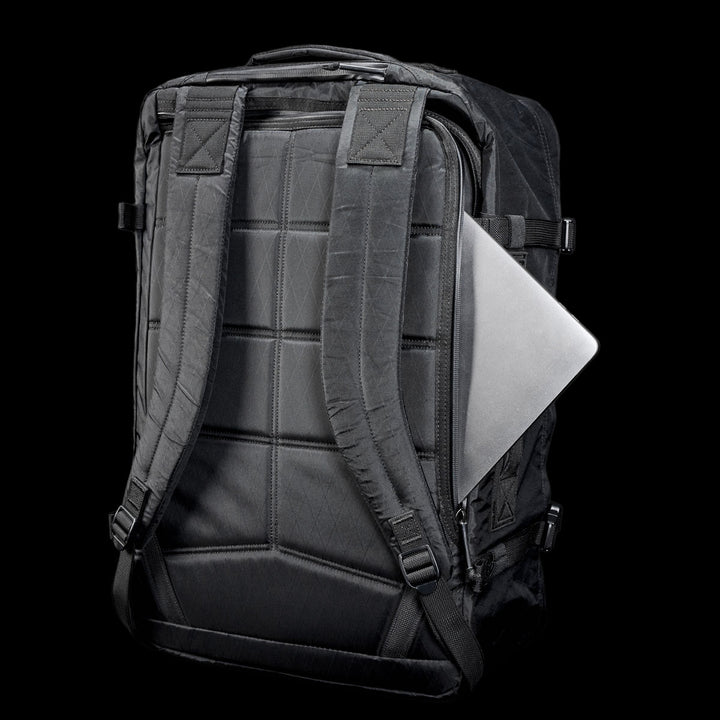 The GR3 - X-PAC, a black waterproof carry-on backpack, features a silver laptop partially tucked into its back pocket, shown against a black background.