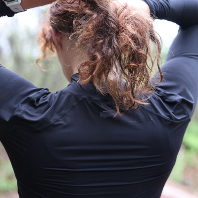 Wearing black Women's Long Sleeve Mock Neck - ToughMesh, a person with curly hair adjusts their ponytail outdoors, enjoying the comfort of featherweight fabric.