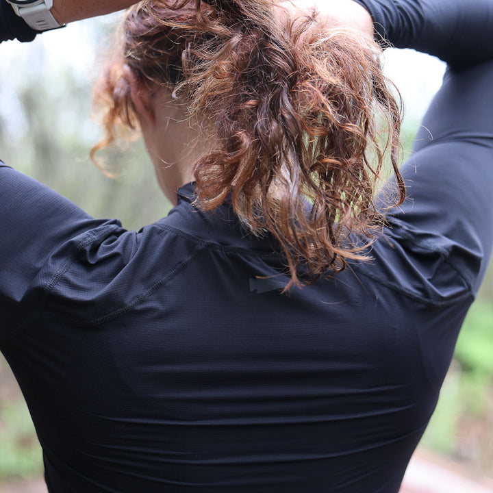 Wearing black Women's Long Sleeve Mock Neck - ToughMesh, a person with curly hair adjusts their ponytail outdoors, enjoying the comfort of featherweight fabric.