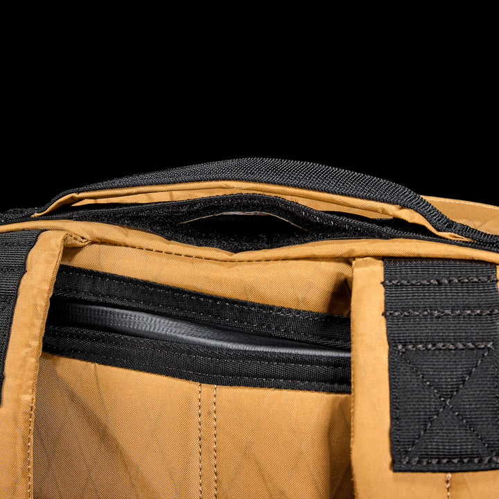 Close-up of the GR0 - X-PAC bag, crafted from waterproof material in brown with black handles and a partially open zipper, set against a black background.