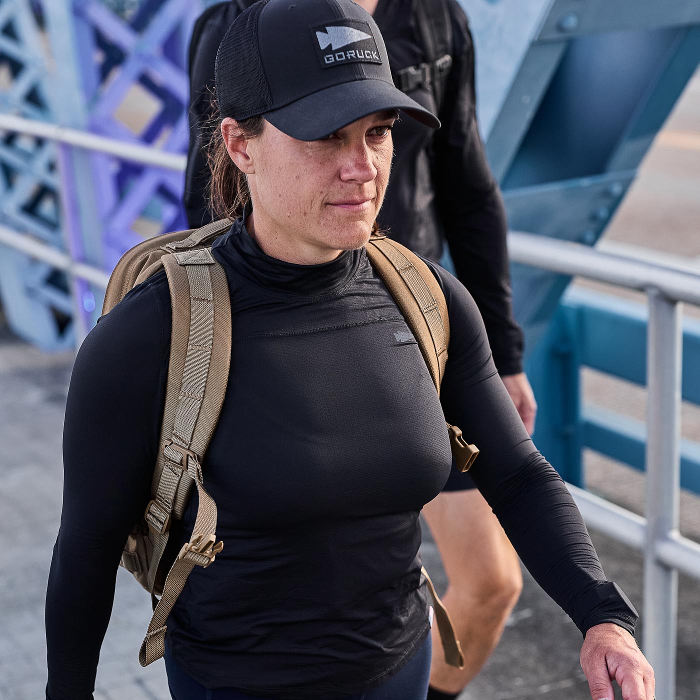 A person wearing a black cap and Women’s Long Sleeve Mock Neck - ToughMesh top, equipped with a tan backpack featuring Italian ToughMesh, walks on a bridge.