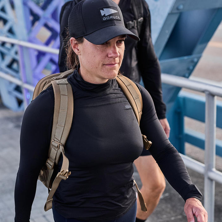 A person wearing a black cap and Women’s Long Sleeve Mock Neck - ToughMesh top, equipped with a tan backpack featuring Italian ToughMesh, walks on a bridge.