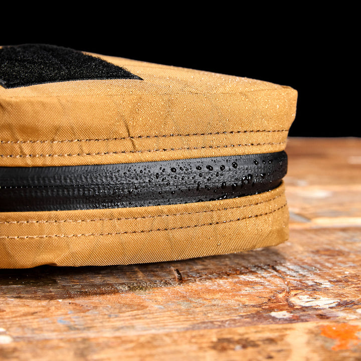 Close-up of the GR1 Field Pocket - X-PAC, a tan electronics organizer made from waterproof X-PAC fabric with a black AquaGuard zipper, showing water droplets on the surface atop a wooden table.
