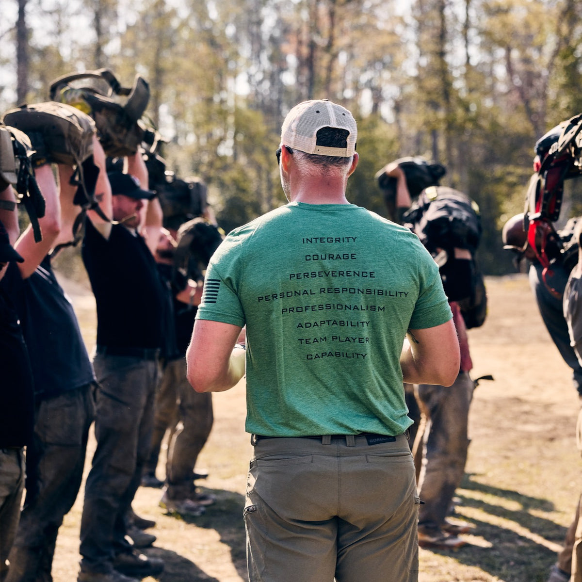 A man in a green t-shirt with a list of values on the back stands before a group wearing abrasion resistant ToughDry pants, all holding rucksacks over their heads during an outdoor workout.
