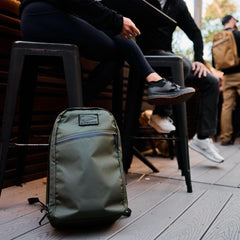 GORUCK olive green durable backpack on wooden floor near people sitting on stools outdoors