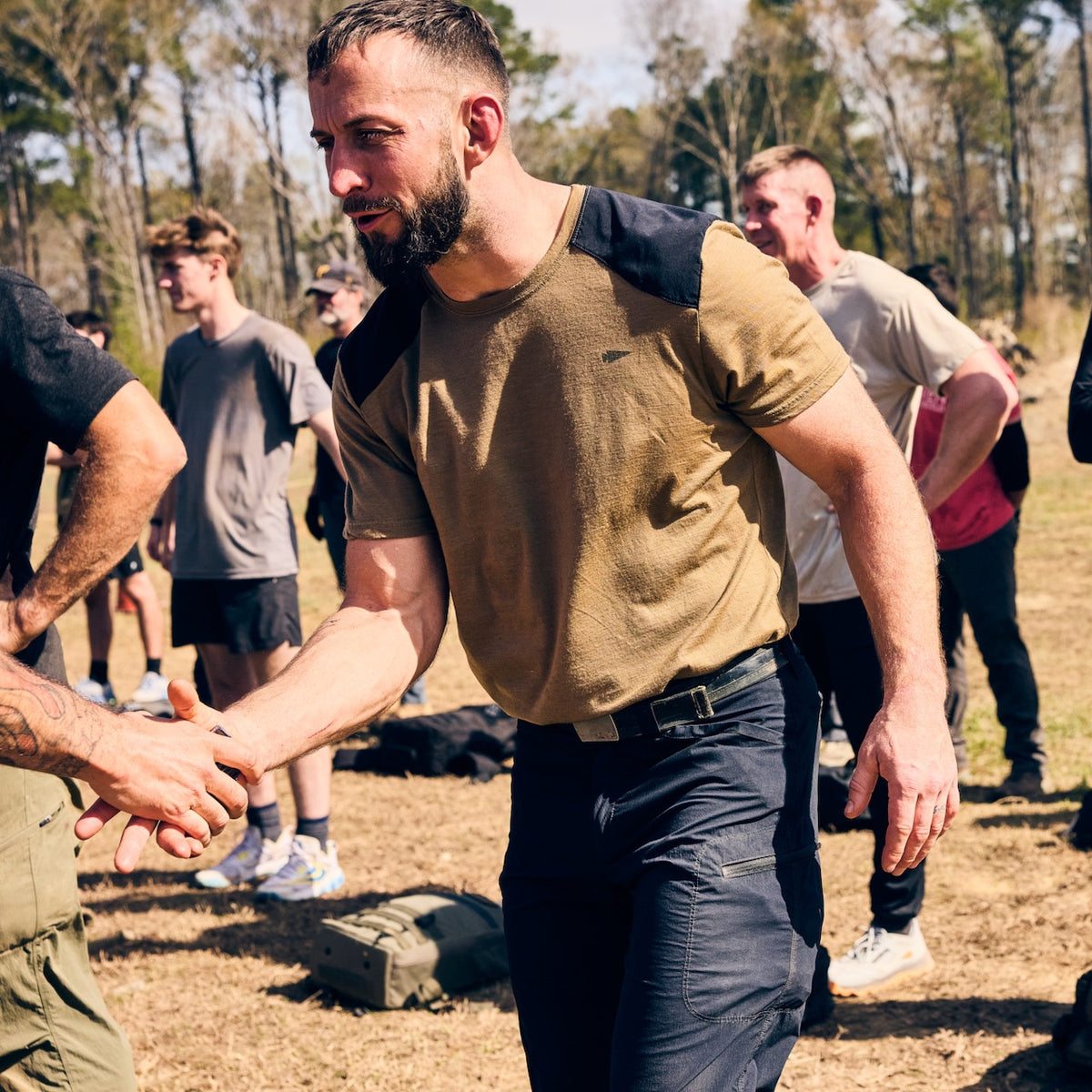 A bearded man in a tan t-shirt and abrasion resistant ToughDry pants shakes hands with another person in a field.