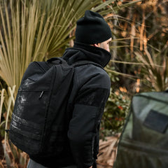 Man wearing black beanie and jacket carrying dusty black GORUCK rucksack outdoors near plants
