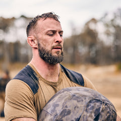A bearded man lifts a heavy sandbag outdoors, wearing a tan shirt and looking focused—developing core strength just like training with the Sand Medicine Ball in his home gym.