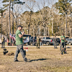 A man in a green t-shirt and ToughDry pants speaks into a microphone while holding papers, addressing a group of people standing in a field in front of parked trucks and trees.