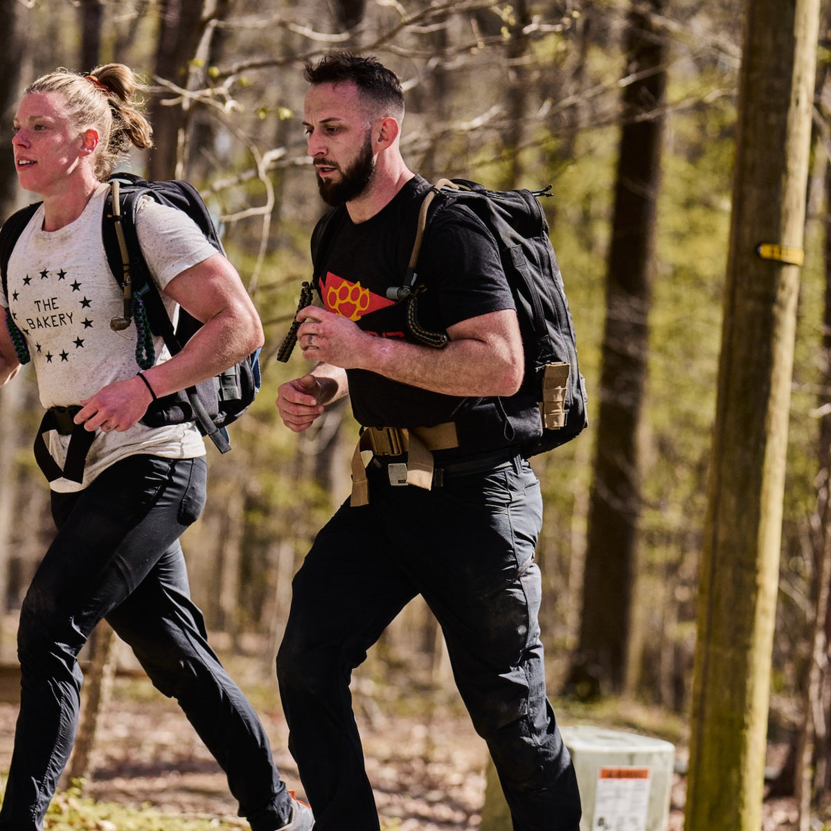 A woman and a man wearing weighted backpacks and ToughDry pants run side-by-side on a wooded trail.