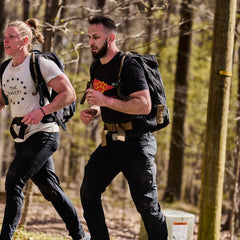 A woman and a man wearing weighted backpacks and ToughDry pants run side-by-side on a wooded trail.