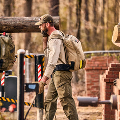 A bearded man in a baseball cap and tactical backpack walks while carrying a large log, wearing abrasion-resistant, fast-drying technical pants.