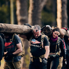 A group of people wearing backpacks and ToughDry pants work together to carry a heavy log on their shoulders through a wooded area.