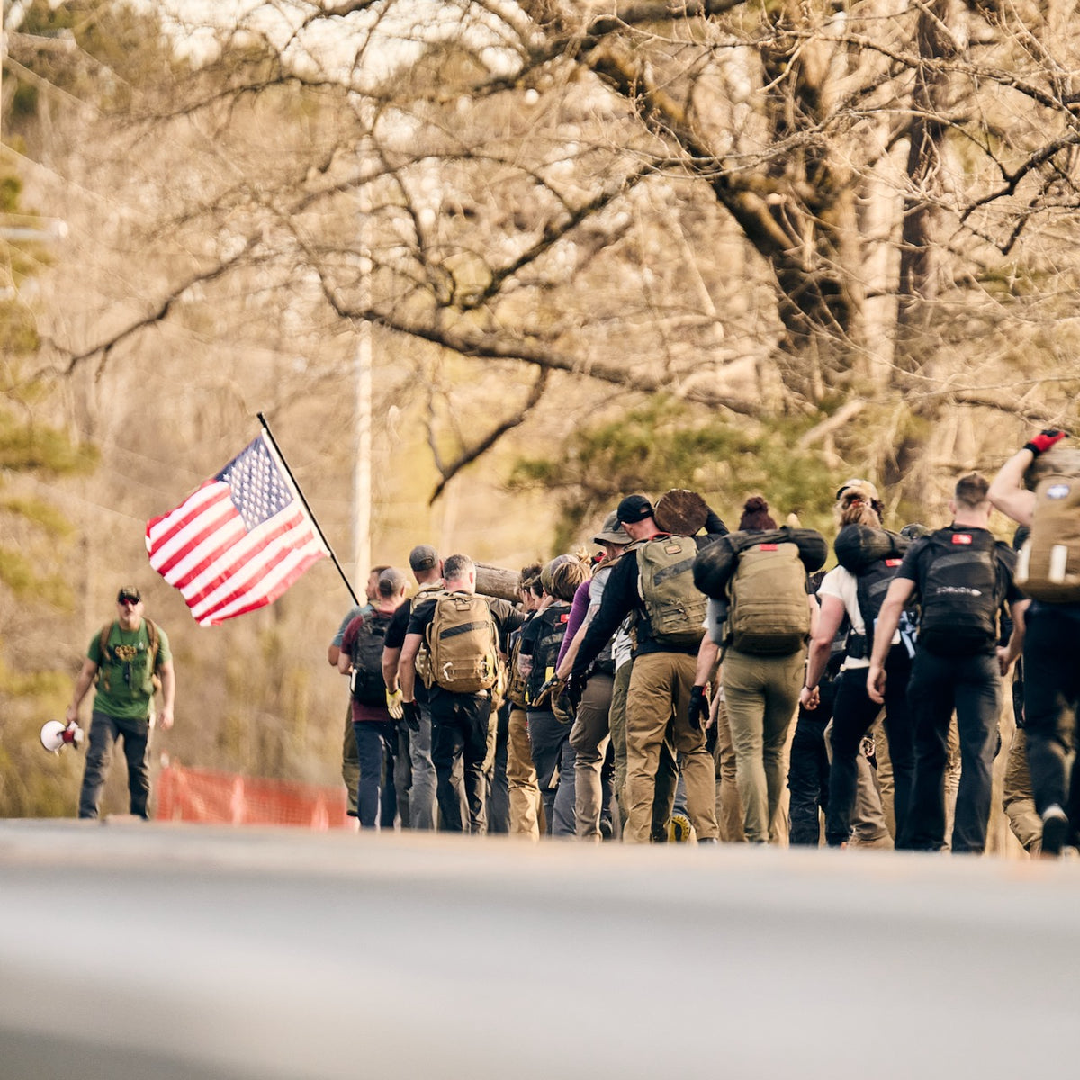 A group of people wearing backpacks and abrasion resistant pants walk together along a road, with one person carrying an American flag.