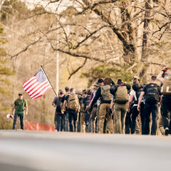A group of people wearing backpacks and abrasion resistant pants walk together along a road, with one person carrying an American flag.