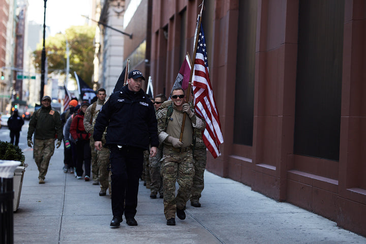 A group of people, some in uniform, walk down a city sidewalk, one carrying an American flag.