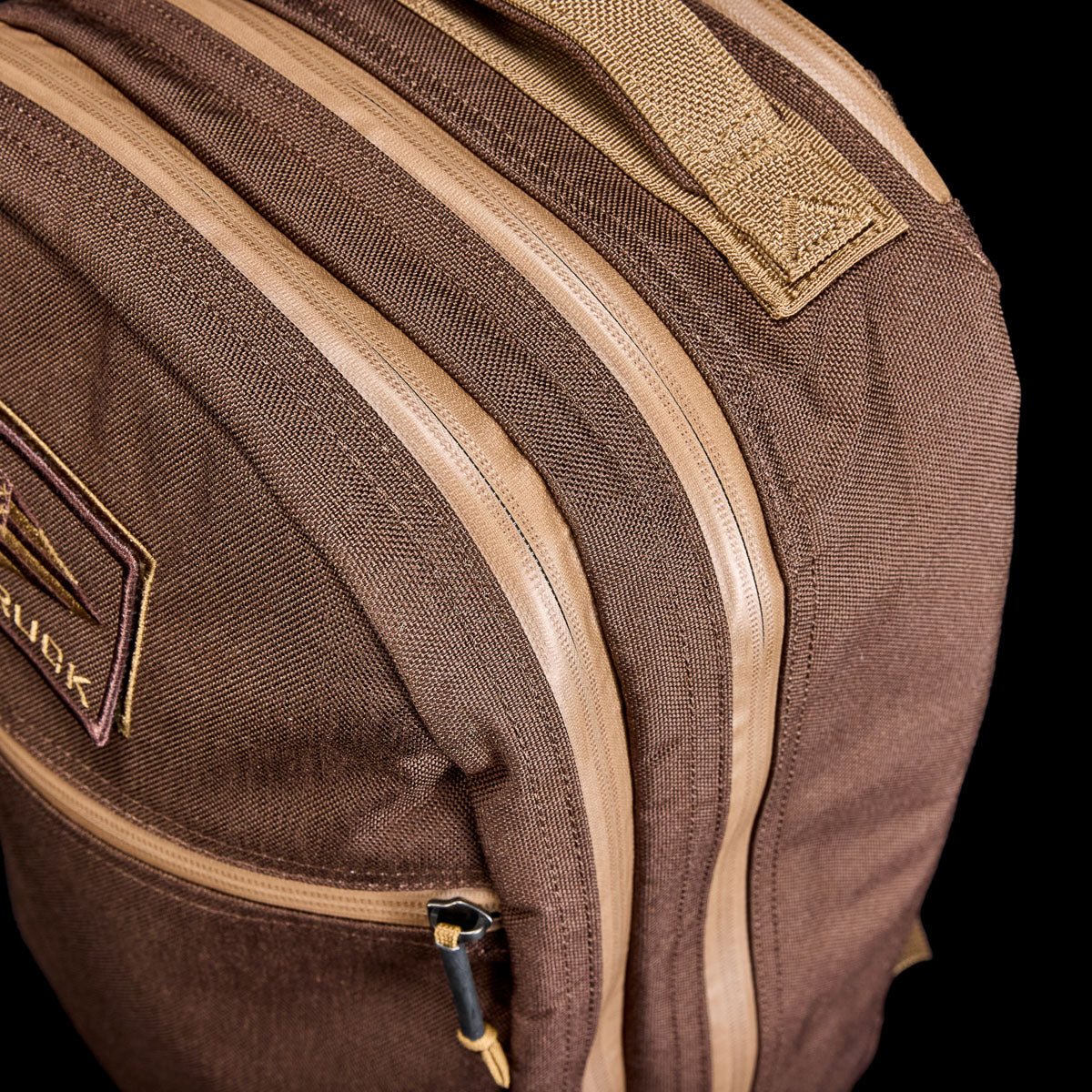 Close-up of the Double Bullet Ruck w/ Bombproof Laptop Compartment - 18L: a brown travel backpack with tan zippers, bombproof laptop storage, and a small patch on the front pocket.