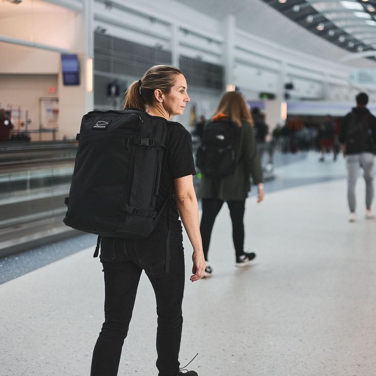 A woman walks through a busy airport terminal with the GR3 - X-PAC carry-on backpack, its waterproof material standing out as other travelers move in the background.