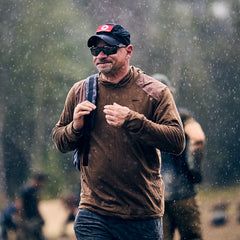 A man in a brown jacket and black cap, wearing the Men’s Commando Pullover - Merino Wool, walks in the rain with a backpack; blurred people are visible in the background.
