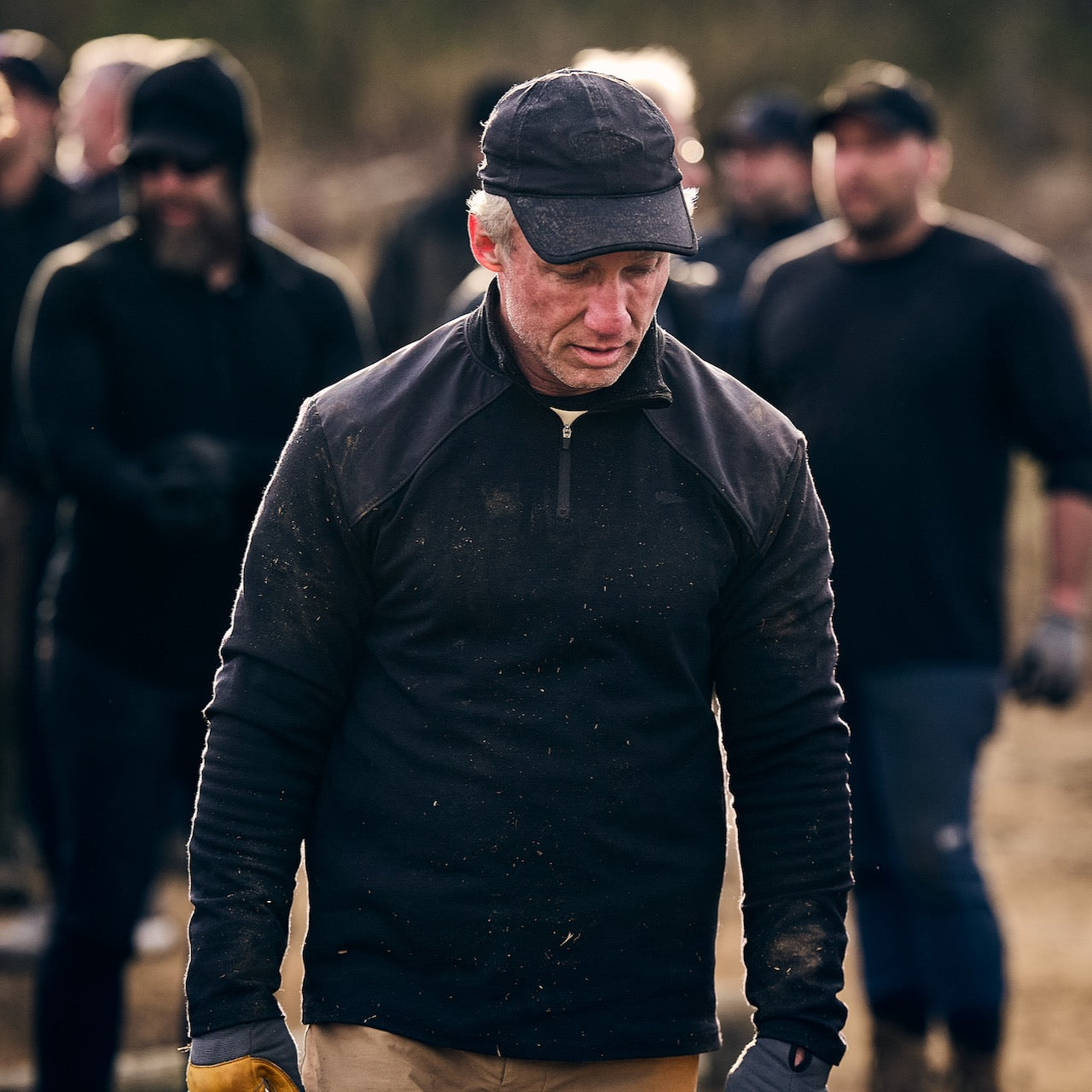 A man in dark clothes and a cap stands outdoors wearing the Men's Heavy Commando Quarter Zip - Merino Wool, which keeps him fresh; a group of people is blurred in the background.
