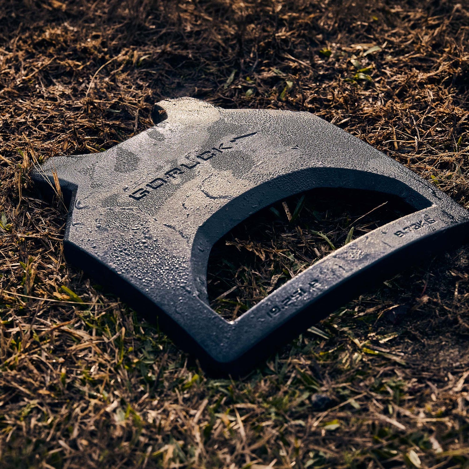 Black Curved Ruck Plate rests on dew-covered grass and dirt, angled to display the brand name and engraved weight—ideal for increasing the challenge in your rucking weight vest.