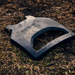 Black Curved Ruck Plate rests on dew-covered grass and dirt, angled to display the brand name and engraved weight—ideal for increasing the challenge in your rucking weight vest.