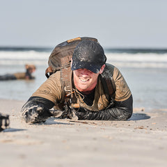 Man in sandy clothes and a Slick Short Sleeve Tee - Tri-BlendX backpack crawling on a beach, smiling, with ocean waves in the background.